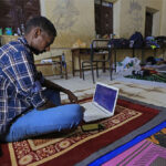 A man uses a laptop computer while sitting on a mat in a classroom