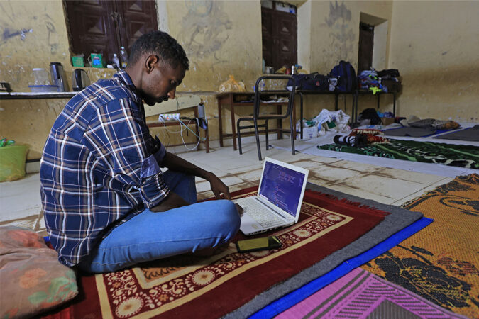 A man uses a laptop computer while sitting on a mat in a classroom