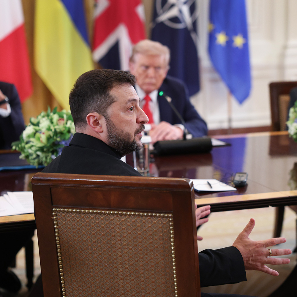 President Volodymyr Zelensky sits at a table, with President Trump in the background