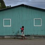 A woman carrying a child walks past a green building in Colombia