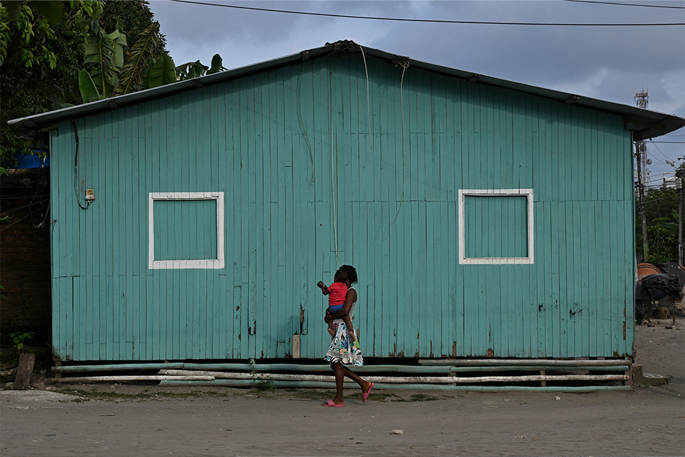 A woman carrying a child walks past a green building in Colombia